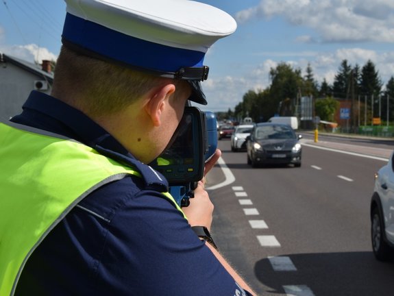 Na zdjęciu umundurowany policjant ruchu drogowego mierzący prędkość poruszających się po drodze pojazdom.