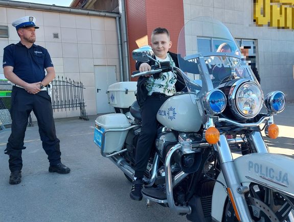 Na zdjęciu chłopiec siedzący na motocyklu służbowym. Obok stoi umundurowany policjant ruchu drogowego.