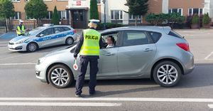 Na zdjęciu umundurowana policjantka ruchu drogowego na jezdni bada stan trzeźwości kierującego srebrnym samochodem. W tle radiowóz policyjny.