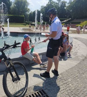 Na zajęciu umundurowany policjant patrolu rowerowego obok fontanny multimedialnej w Rzeszowie na al. Lubomirskich, obok niego chłopiec, któremu policjant daje ulotkę.