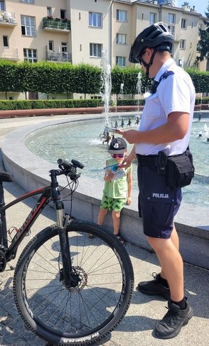 Na zajęciu umundurowany policjant patrolu rowerowego obok fontanny multimedialnej w Rzeszowie na al. Lubomirskich, obok niego chłopiec, któremu policjant daje ulotkę.