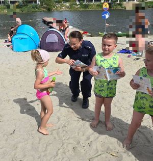 Na zdjęciu umundurowana policjantka na plaży miejskiego kąpieliska Żwirownia przy ul. Kwiatkowskiego, obok niej dwoje małych chłopców. W tle zbiornik wodny i bloki.