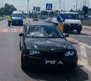Na zdjęciu pojazd marki BMW z widocznym pęknięciem szyby czołowej po stronie prawej. W tle policyjny ambulans ruchu drogowego i umundurowani policjanci. Z boku inne pojazdy.