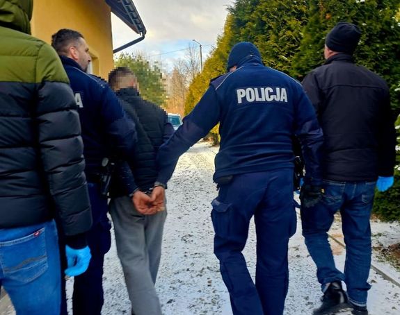 Na zdjęciu umundurowani policjanci prowadzą mężczyznę, który na ręce trzymane z tyłu ma założone kajdanki. Obok policjanci w ubraniach cywilnych.