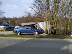 Na zdjęciu samochód marki opel koloru niebieskiego, który wjechał w metalowy garaż. Po prawej stronie oraz w tle rosnące drzewa. Z lewej strony ułożone drzewo kominkowe przykryte niebieską folią. W tle fragment budynku.