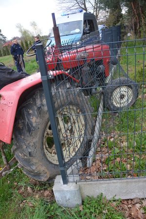 Na zdjęciu ciągnik samodzielnej konstrukcji koloru czerwonego, który prawym bokiem wjechał w ogrodzenie posesji. W tyle radiowozu oraz policjanci pracujący na miejscu zdarzenia