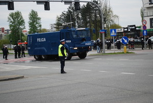 Na zdjęciu na środku skrzyżowania umundurowany policjant ruchu drogowego, który ręcznie steruje ruchem. W  dalszym planie policyjna polewaczka, umundurowani policjanci oraz przechodnie. W dalszym tel budynki mieszkalne.