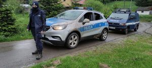 Na zdjęciu umundurowany policjant, za nim dwa radiowozu. W tle drzewa i domy.