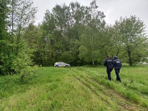 Na zdjęciu dwóch umundurowanych policjantów idących w kierunku lasu. W tle radiowóz policyjny i las.