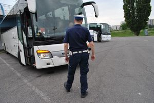 Na zdjęciu, na parkingu stojący tyłem w umundurowaniu służbowym policjant ruchu drogowego. Policjant stoi przodem do autokaru w kolorze białym. Z prawej strony, na dalszym planie drugi, taki sam autokar.