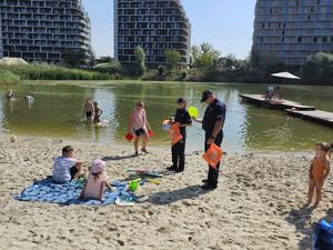 Na zdjęciu dwoje umundurowanych policjantów w granatowych mundurach, rozmawiają z wypoczywającymi zad miejskim kąpieliskiem Żwirownia. Za nimi kąpielisko, w tle wieżowce.