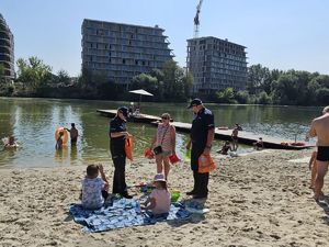 Na zdjęciu dwoje umundurowanych policjantów w granatowych mundurach, rozmawiają z wypoczywającymi zad miejskim kąpieliskiem Żwirownia. Za nimi kąpielisko, w tle wieżowce.