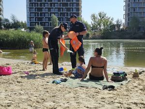 Na zdjęciu dwoje umundurowanych policjantów w granatowych mundurach, rozmawiają z wypoczywającymi zad miejskim kąpieliskiem Żwirownia. Za nimi kąpielisko, w tle wieżowce.