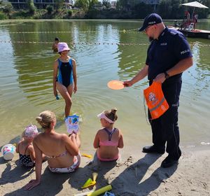 Na zdjęciu umundurowany policjant w granatowym mundurze rozmawia z wypoczywającymi zad miejskim kąpieliskiem Żwirownia. Za nim kąpielisko, w tle wieżowce.