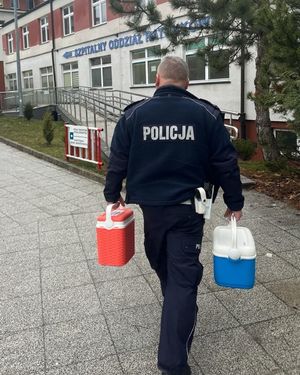 Na zdjęciu idący tyłem, w kierunku Szpitalnego Oddziału Ratunkowego umundurowany policjant ruchu drogowego. W prawej ręce trzyma pojemnik koloru niebieskiego, w lewej czerwonego.