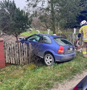 Na zdjęciu pojazd honda, który zawisł na drewnianym płocie. Z prawej strony strażak.