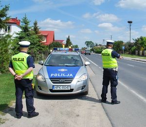 Na zdjęciu dwóch umundurowanych policjantów ruchu drogowego. Jeden z nich stoi na poboczu, drugi używa urządzenia kontrolno-pomiarowego. Przed mini radiowóz policyjny. W tle jadące drogą samochodu. Wzdłuż drogi domy.