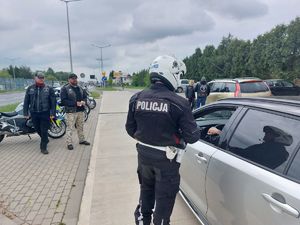 Na zdjęciu umundurowany policjant ruchu drogowego-motocyklista, podczas kontroli drogowej. W tle członkowie klubu motocyklowego.