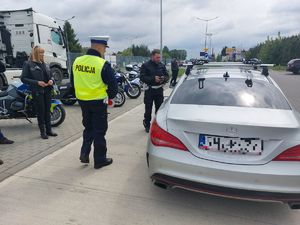 Na zdjęciu umundurowany policjant ruchu drogowego podczas kontroli drogowej. W tle członkowie klubu motocyklowego.