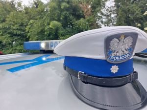 Na zdjęciu policyjna czapka z biały otokiem, za nią belka policyjnego radiowozu z napisem policji. W tle drzewa.