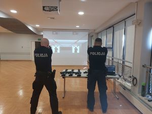 Na zdjęciu dwóch umundurowanych policjantów w granatowych mundurach. Policjanci stoją tyłem, zwróceni są w kierunku multimedialnej tarczy, w rękach trzymają repliki broni.