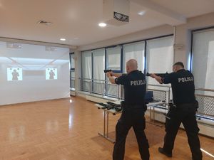 Na zdjęciu dwóch umundurowanych policjantów w granatowych mundurach. Policjanci stoją tyłem, zwróceni są w kierunku multimedialnej tarczy, w rękach trzymają repliki broni.