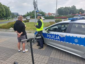 Na zdjęciu dwie umundurowane policjantki ruchu drogowego oraz odwrócony plecami mężczyzna. Obok, po lewej stronie hulajnoga,  po prawej policyjny radiowóz.