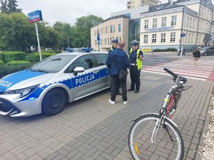 Na zdjęciu dwie umundurowane policjantki ruchu drogowego oraz odwrócony plecami mężczyzna. Obok, po lewej stronie policyjny radiowóz, po prawej rower.