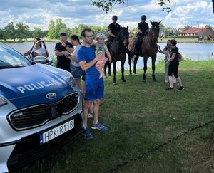 Na zdjęciu policyjny radiowóz. w Tle partol konny przy którym stoją dzieci i dorośli.