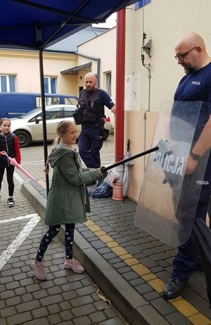 Na zdjęciu umundurowany policjant trzyma tarczę, przed nim dziewczynka z pałką służbową.