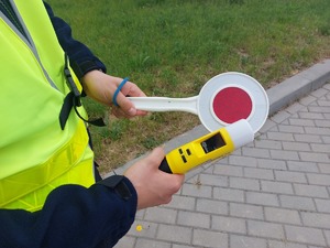 Na zdjęciu policjant w żółtej kamizelce odblaskowej trzyma w ręce urządzenie do badania stanu trzeźwości i tarczę do zatrzymywania pojazdów.