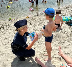 Na zdjęciu umundurowana policjantka wręcza chłopcu ulotkę. W tle woda.