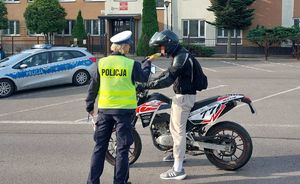 Na zdjęciu umundurowana policjantka ruchu drogowego stoi na środku jezdni. Na drugim pasie w lewo stoi motocykl. Policjantka kontroluje stan trzeźwości motocyklisty. W tle radiowóz policyjny i drugi umundurowany policjant ruchu drogowego.