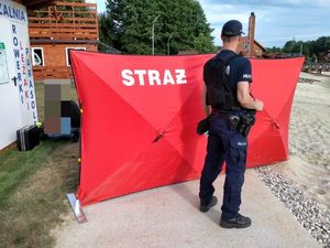 Na zdjęciu umundurowany policjant na tle parawanu z napisem straż. W tle drzewa i budynek.