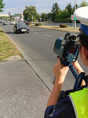 Na zdjęciu umundurowana policjanta ruchu drogowego z ręcznym miernikiem prędkości, w tle jezdnia.