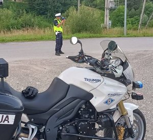 Na zdjęciu umundurowany policjant ruchu drogowego z ręcznym miernikiem prędkości, obok policyjny motocykl.