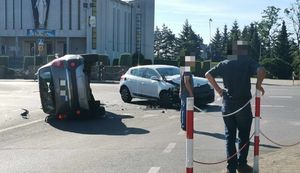 Na zdjeciu skrzyżowanie, na jezdni ba doku leży pojazd marki Ople,  obok niego z prawej strony stoi rozbity pojazd marki Renault. Przed samochodami stoją odwróceniu tyłem mężczyźni. W tle kościół.