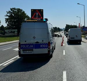 Na zdjęciu policyjny ambulans ruchu drogowego, ustawiony na jezdni z włączoną tablicą świetlną informującą o wypadku. Przed radiowozem, na jezdni stoi motocykl. W tel budynki i inne pojazdy.