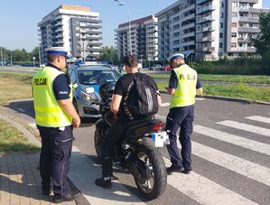 Na zdjęciu dwoje umundurowanych policjantów ruchu drogowego z żółtych kamizelkach z napisem policja na plecach, prowadzą kontrolę kierującego motocyklem. W tel policyjny radiowóz.