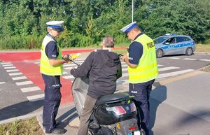 Na zdjęciu dwoje umundurowanych policjantów ruchu drogowego z żółtych kamizelkach z napisem policja na plecach, prowadzą kontrolę kierującego motocyklem. W tel policyjny radiowóz.