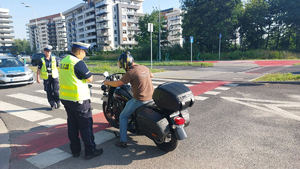 Na zdjęciu dwoje umundurowanych policjantów ruchu drogowego z żółtych kamizelkach z napisem policja na plecach, prowadzą kontrolę kierującego motocyklem. W tel policyjny radiowóz.