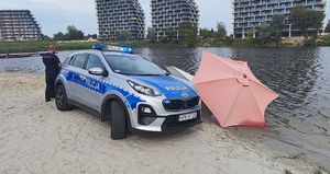 Na zdjęciu policyjny radiowóz, z jego lewej strony odwrócony tyłem stoi umundurowany policjant. Z prawej strony radiowozu leżąsy na ziemi, rozłożony parasol plażowy. W tel woda i wieżowce.