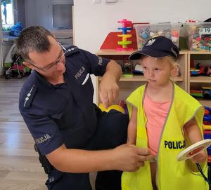 Na zdjęciu, z lewej strony umundurowany policjant, który pomaga ubrać stojącej obok dziewczynce kamizelkę odblaskową. Dziewczynka w ręce trzyma tarczę do zatrzymywania pojazdów. Na łowie ma założoną czapkę policyjną.