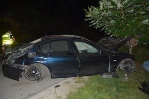 Na zdjęciu uszkodzony pojazd marki bmw, który wjechał w drzewo. Z lewej strony umundurowany policjant w kamizelce odblaskowej.