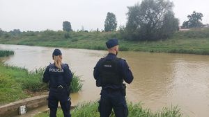 Na zdjęciu stoi odwróconych tyłem dwoje umundurowanych funkcjonariuszy policji, przed nimi rzeka.