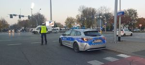 Na zdjęciu umundurowany policjant ruchu drogowego na skrzyżowaniu kieruje ruchem, obok policyjny radiowóz. W tle przejeżdżające pojazdy i przechodnie.