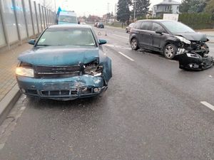 Na jezdni dwa pojazdy jakie brały udział w zdarzeniu drogowym, za nimi policyjny ambulans ruchu drogowego, w tle inne przejeżdżające pojazdy.