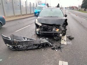 Na jezdni uszkodzony pojazd marki peugeot jaki brały udział w zdarzeniu drogowym, za nim policyjny ambulans ruchu drogowego, w tle inne przejeżdżające pojazdy.