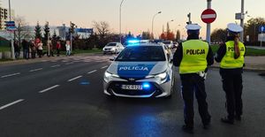 Na zdjęciu dwoje umundurowanych policjantów w odblaskowych kamizelkach stojących na skrzyżowaniu, obok nich policyjny radiowóz z włączoną belką. Po lewej stronie przed przejściem dla pieszych stoją piesi.