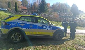 Na zdjęciu dwoje policjantów, obok nich radiowóz. W tle cmentarz.